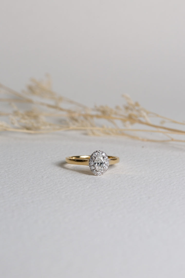 Diamond engagement ring set in 18ct gold.  An oval-cut diamond is at the centre, with a halo of diamonds around it
