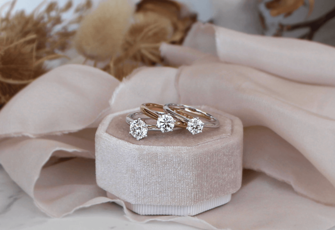 Three solitaire diamond engagement rings, set with 0.50ct 0.70ct and 1.00ct natural diamonds, are placed on a pink velvet pad.