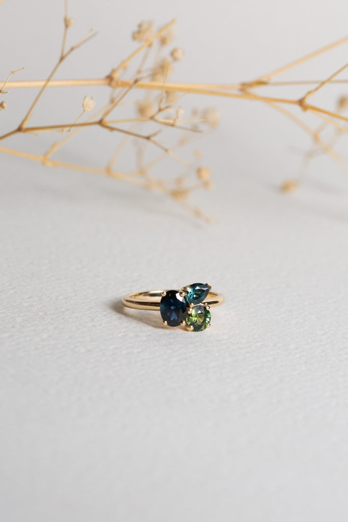 A Sapphire ring set with three different Sapphires.  There is an oval Blue Sapphire, a pear Teal Sapphire, and a round Green Sapphire.