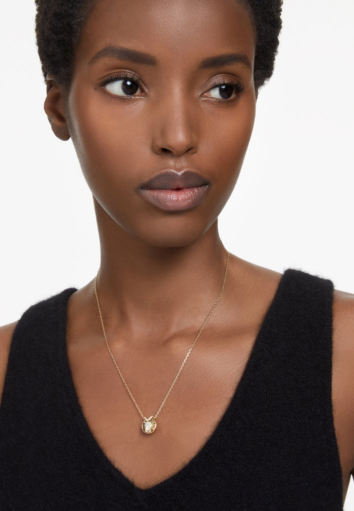 A model is wearing a gold Swarovski Bella pendant on a chain