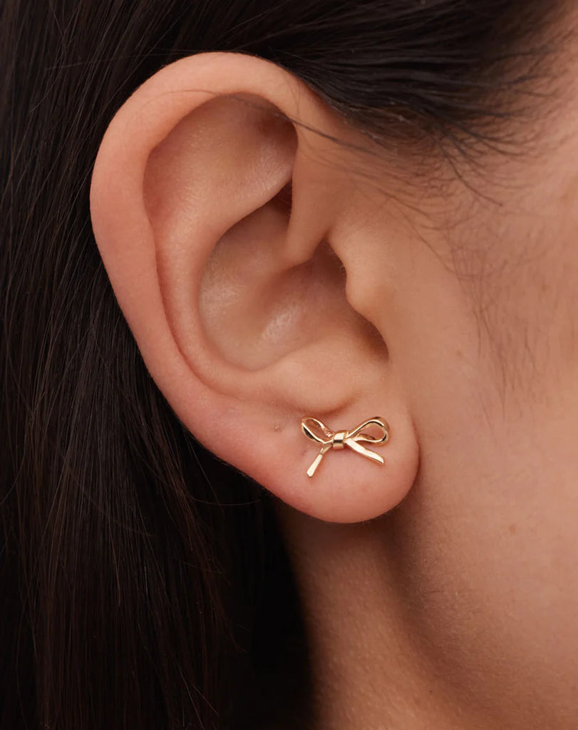 Small gold bow stud earrings shown on the ear