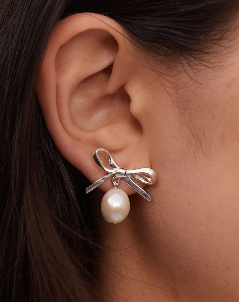 A model wears an oversized silver bow earring with a pearl hanging off them